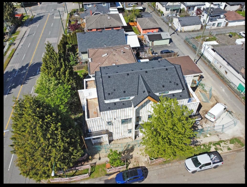Victoria Haus – A high-angle drone view captures a multi-story residential building under construction, wrapped in white weather barrier sheeting beneath a completed dark shingled roof. The property is set within an established urban residential neighborhood bordered by mature green trees, paved streets, and neighboring single-family homes.