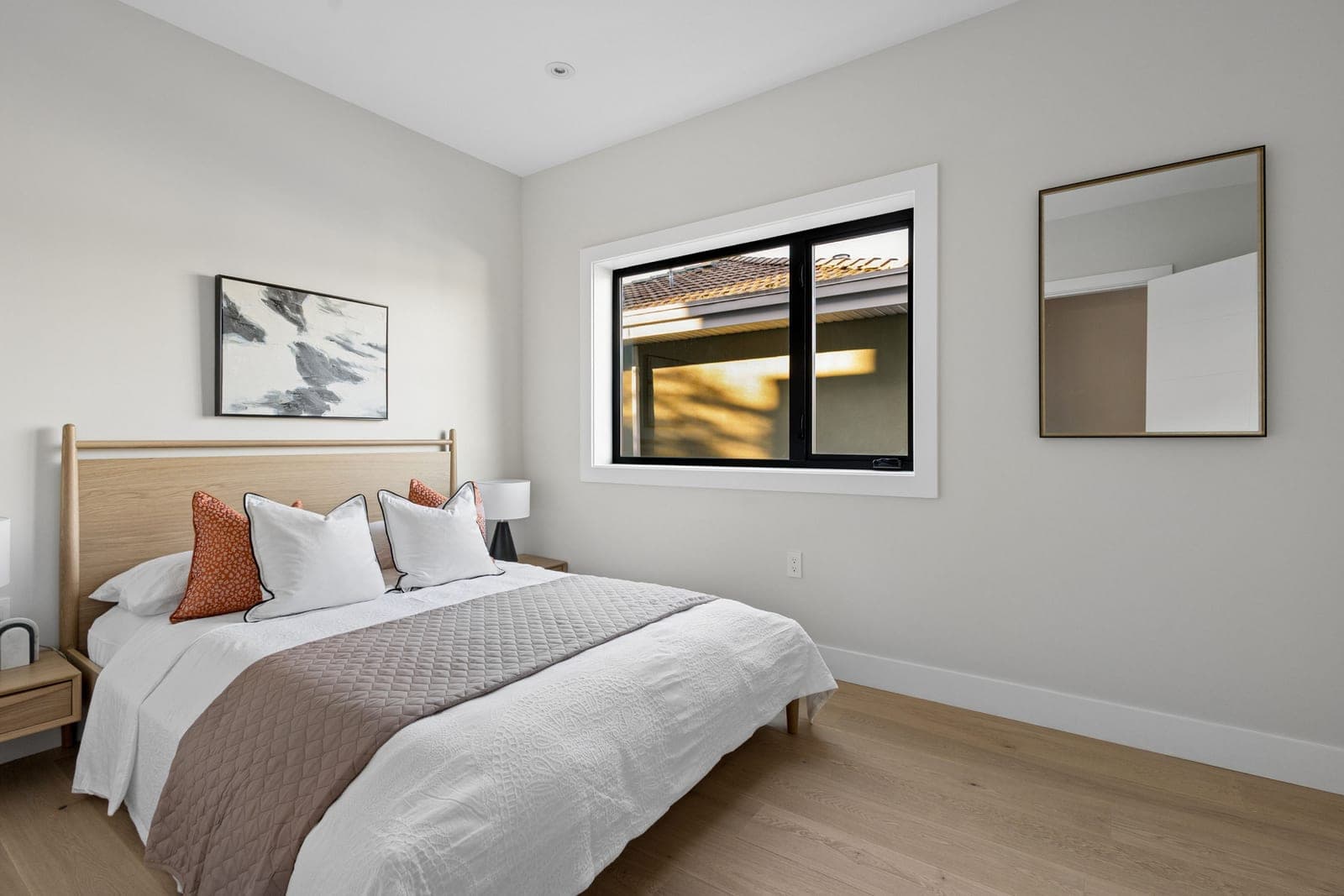 Victoria Haus – A bright, minimally staged bedroom features a light wood spindle-accented bed dressed in crisp white and neutral-toned linens, complemented by light hardwood floors. A black-framed window lets in natural daylight alongside light gray walls decorated with a framed abstract print and a brass-trimmed mirror.