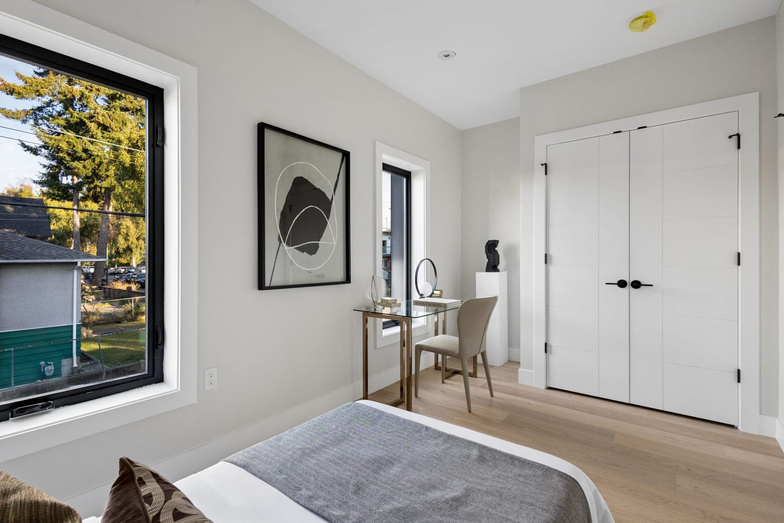 Victoria Haus – A modern, naturally lit bedroom featuring light wood flooring, a glass-topped writing desk with a contemporary chair, and a double-door closet with black hardware. Black-framed windows bring in daylight and offer views of surrounding green trees and neighboring houses.