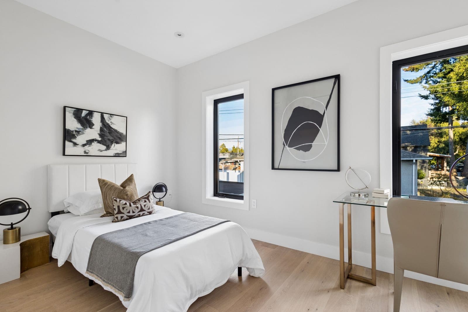 Victoria Haus – A bright, minimalist modern bedroom features a twin bed with a white upholstered headboard, crisp white linens, and decorative accent pillows over light hardwood floors. Two black-framed windows bring in ample daylight, alongside abstract wall art and a small glass-topped desk with a contemporary chair positioned in the corner.