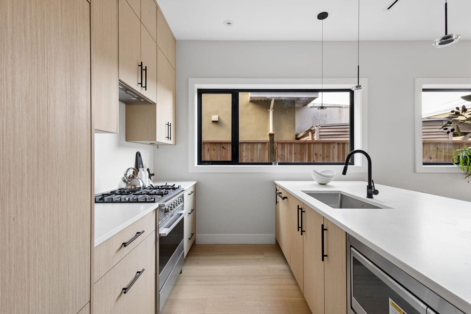 Victoria Haus – A modern, bright galley-style kitchen featuring light wood cabinetry with sleek black hardware, white quartz countertops, and stainless steel appliances. A kitchen island contains an undermount sink with a matte black faucet, set against a backdrop of light hardwood floors and a black-framed window looking out to a wooden fence.