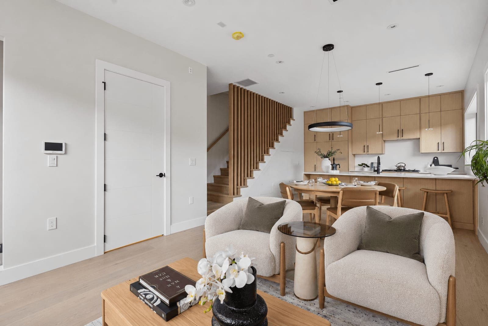 Victoria Haus – An open-concept living, dining, and kitchen area featuring two light-colored bouclé armchairs in the foreground and light wood flooring throughout. In the background, a wooden dining table sits beside a modern kitchen with flat-panel wood cabinetry, white countertops, and a staircase framed by vertical wooden slat railings.