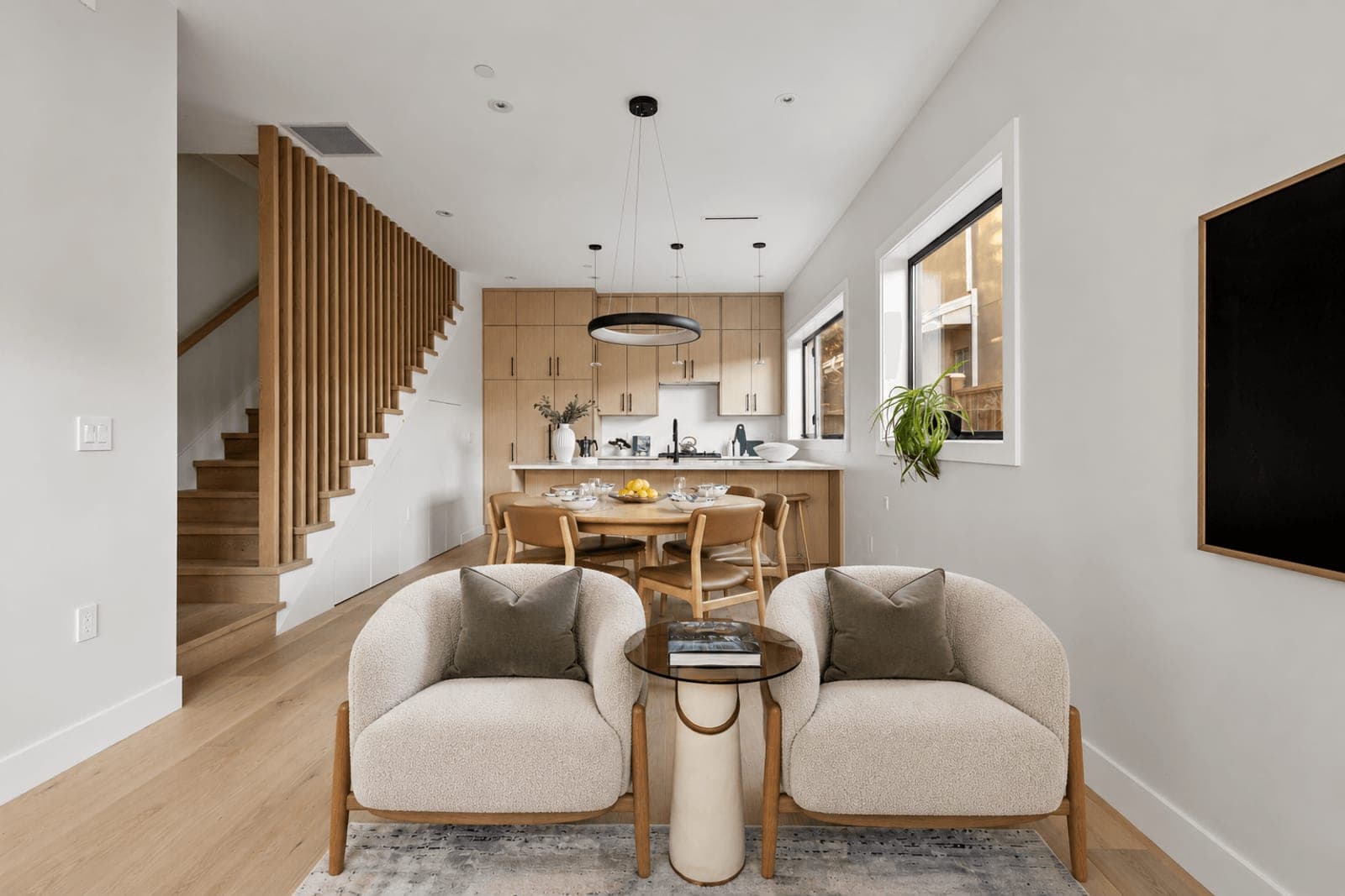 Victoria Haus – A bright, open-concept main floor features a seating area with two cream bouclé armchairs in the foreground, leading to a round wooden dining table and a kitchen with light wood cabinetry. Vertical wooden slats frame a staircase on the left, while white walls, light hardwood floors, and modern minimalist lighting enhance the airy ambiance.
