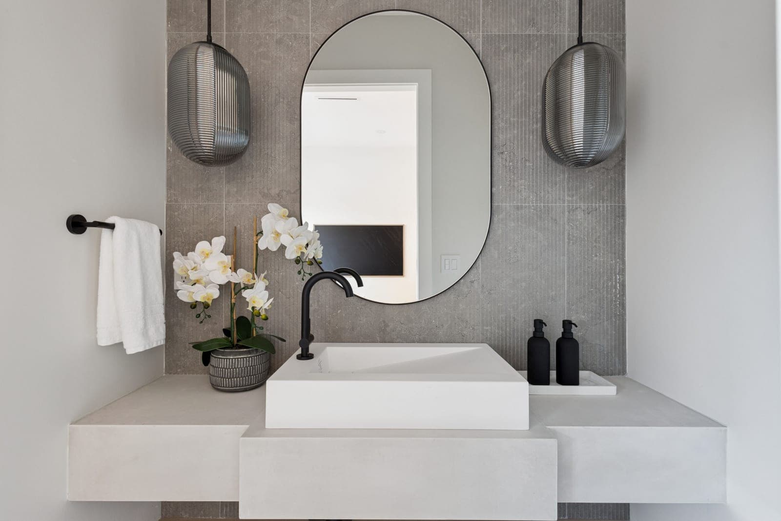 Victoria Haus – A modern bathroom vanity area featuring a rectangular vessel sink with matte black hardware set against a textured grey tile accent wall. An oval mirror hangs centered between two ribbed metallic pendant lights, complemented by white potted orchids and black soap dispensers on the counter.