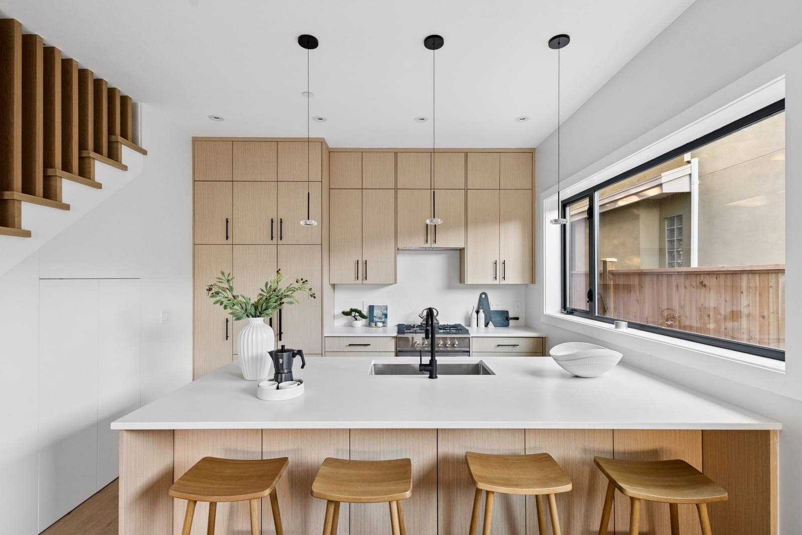 Victoria Haus – A bright, modern kitchen features light wood flat-panel cabinetry, white quartz countertops, and an undermount sink on a spacious island with four wooden stools. Daylight streams in from a wide side window, while black pendant lights, a black faucet, and an adjacent wood-slat staircase complement the clean, minimalist design.