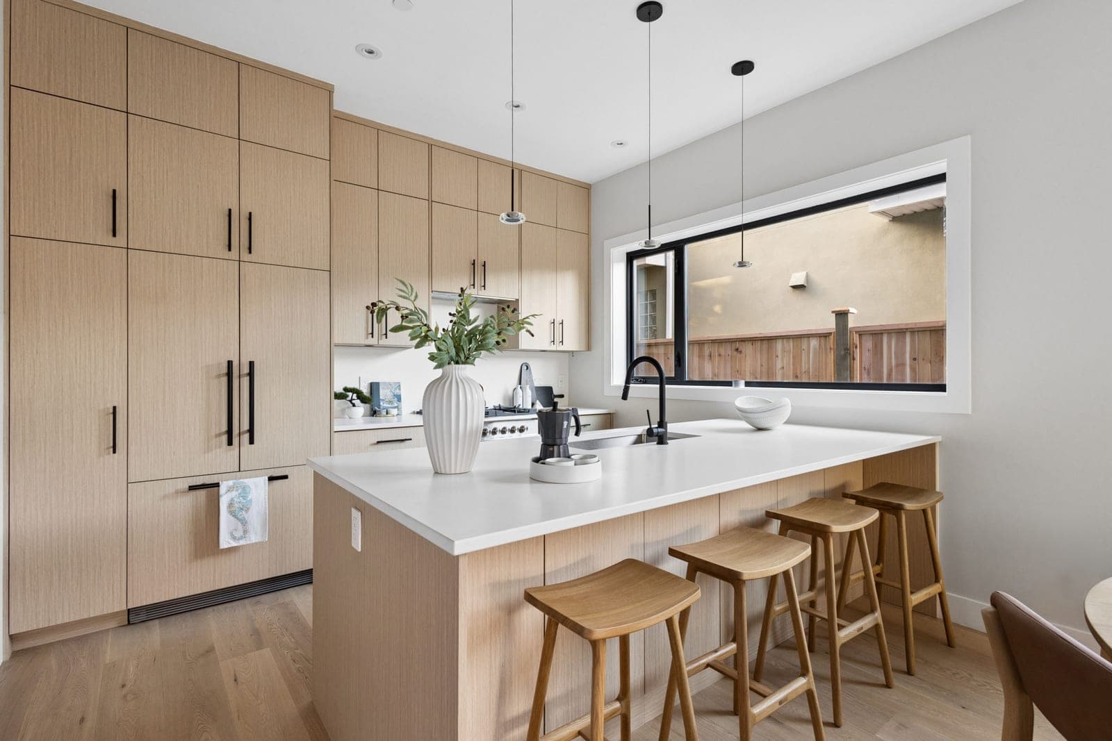 Victoria Haus – A modern kitchen featuring light natural wood flat-panel cabinetry, a spacious white quartz island, and matching wooden barstools. A large horizontal window behind the sink looks out onto a wooden privacy fence, letting in bright natural light that complements the sleek matte black fixtures.