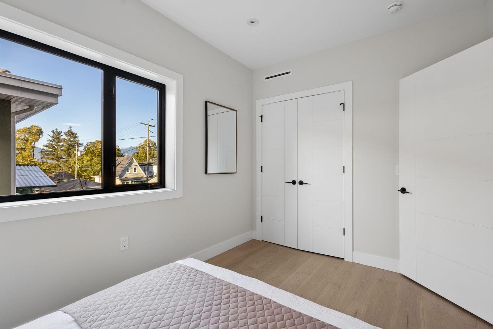 Victoria Haus – A bright, minimalist bedroom featuring light wood flooring, white walls, and a quilted bedspread in the foreground. A large black-framed window offers views of surrounding neighborhood rooftops and distant mountains under a clear blue sky.