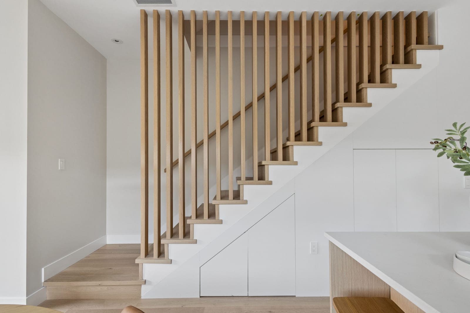 Victoria Haus – A modern interior featuring a light wood staircase framed by full-height vertical wood slats acting as a screen and railing. Built-in white storage cabinetry is seamlessly tucked beneath the stairs, complemented by matching light hardwood floors and crisp white walls.