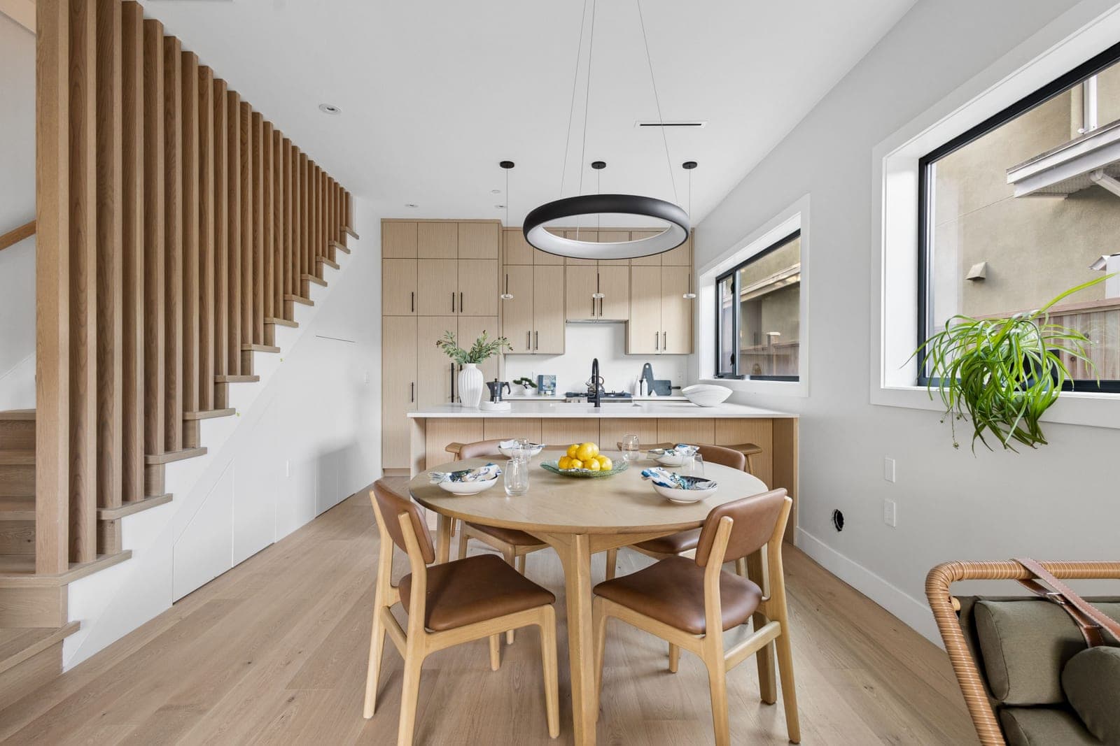 Victoria Haus – A modern open-concept kitchen and dining area features a round wooden dining table surrounded by leather-cushioned chairs beneath a large circular halo pendant light. Natural wood tones dominate the space, seen in the light hardwood flooring, flat-panel kitchen cabinetry, and an open staircase with vertical wood slat railings.