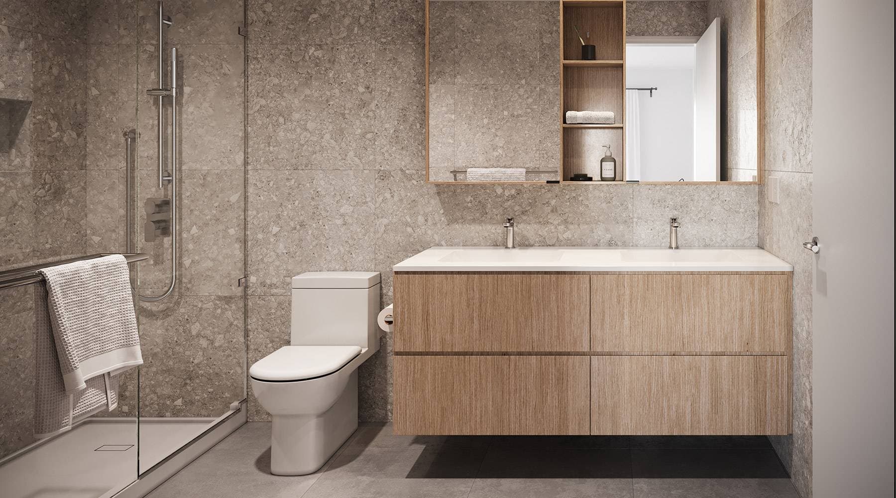 FORM – A modern ensuite bathroom featuring a floating light-wood double vanity and a glass-enclosed walk-in shower. Gray terrazzo-style floor and wall tiles create a sleek, cohesive finish lit by soft interior lighting.