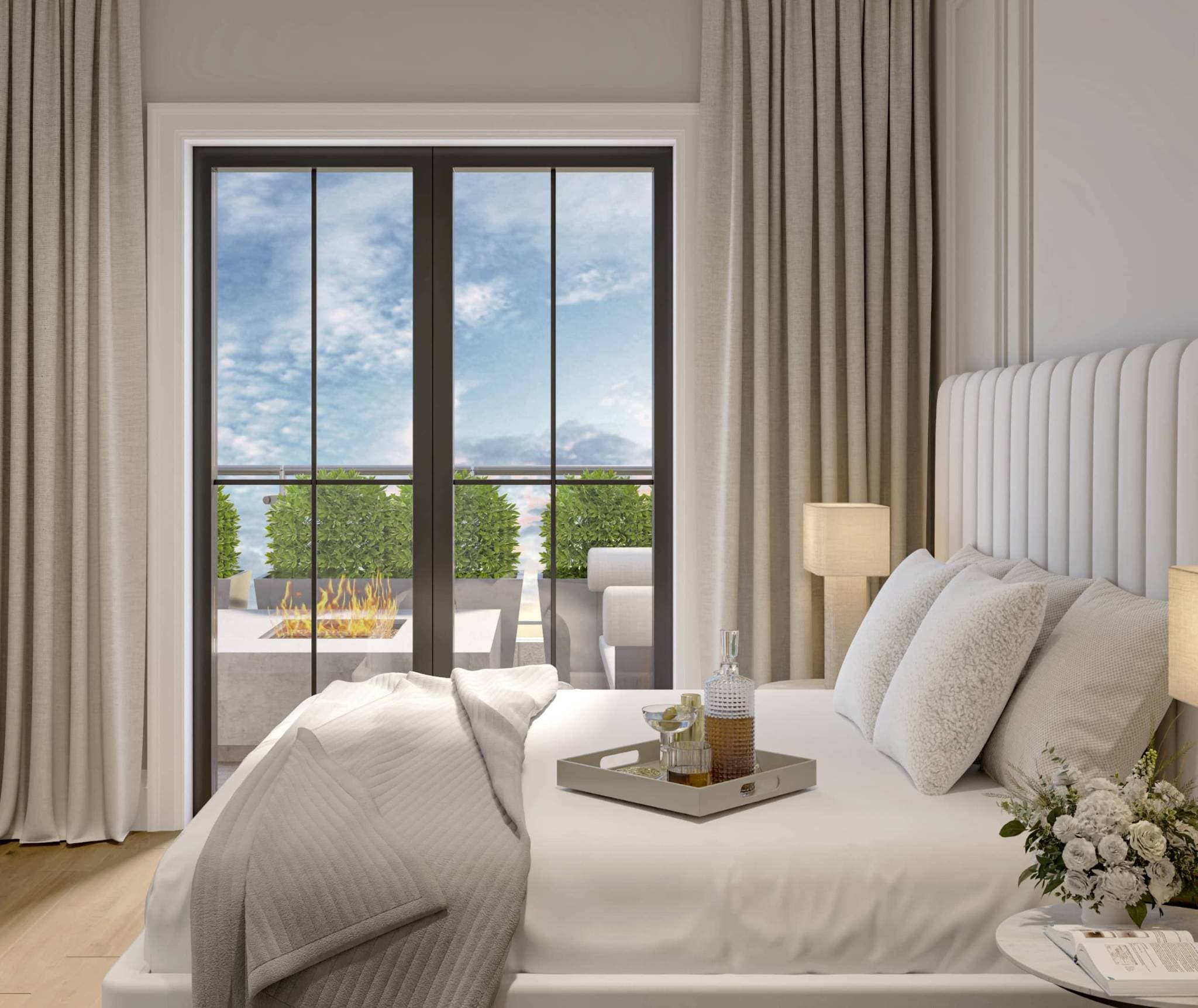 Claremont – A bright, elegant bedroom features a bed dressed in white linens and a channel-tufted headboard, centered around large glass doors. Through the doorway, an outdoor terrace is visible with manicured planters, a glowing fire pit, and a daytime sky.
