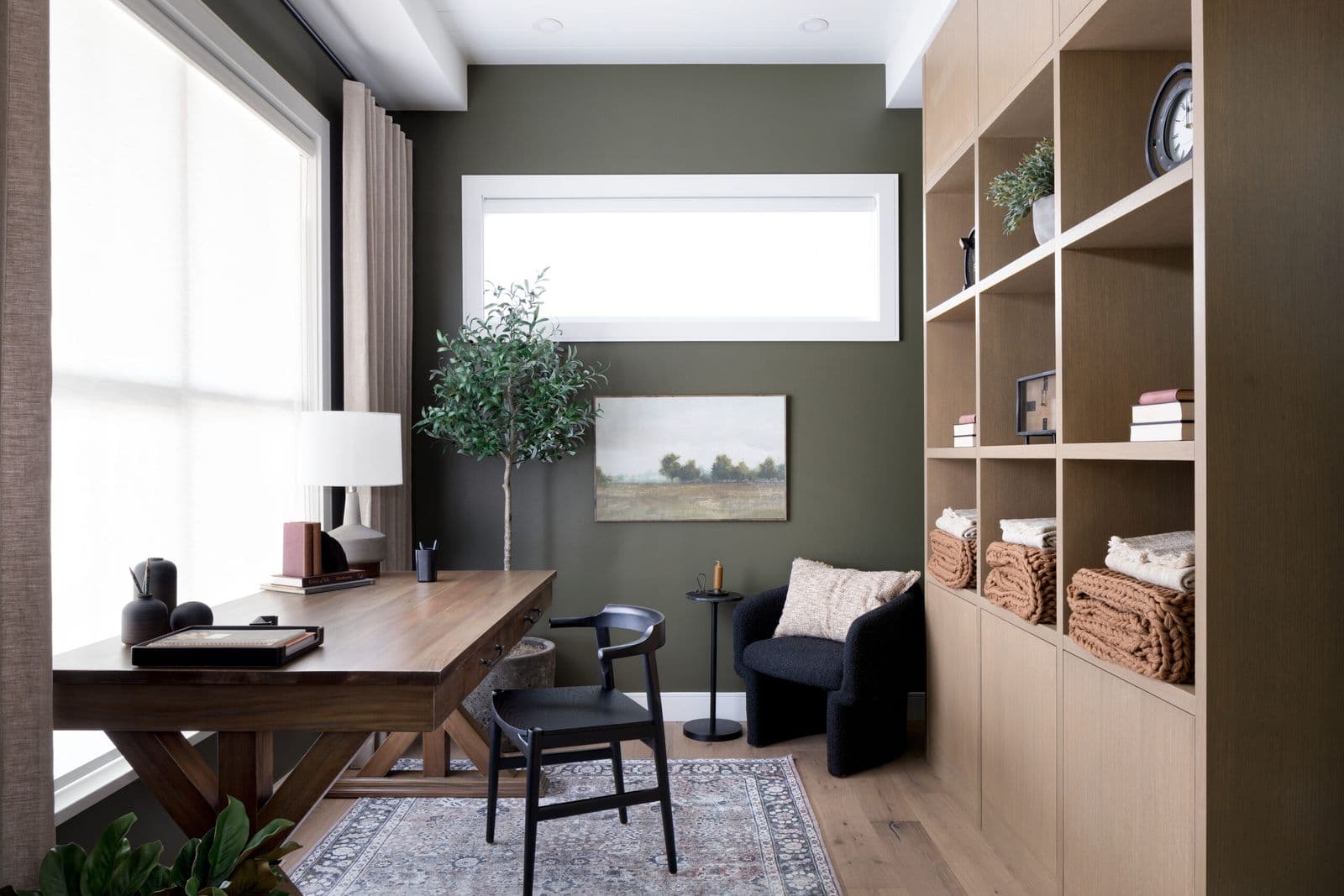 Mirada Estates – Home office with a wood desk, black accent chair, dark green walls, a picture window, and built-in shelving stocked with folded throws and books.