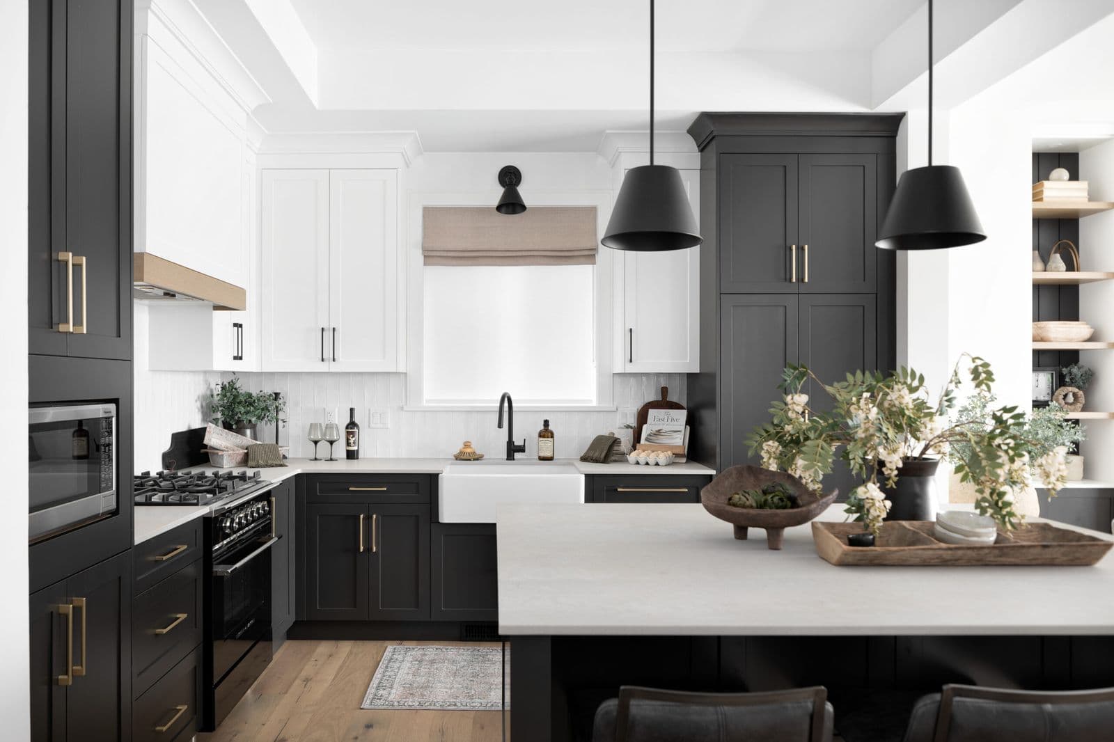 Mirada Estates – Kitchen with black lower cabinets, white uppers, brass hardware, pendant lighting, and an island opening to built-in shelving.