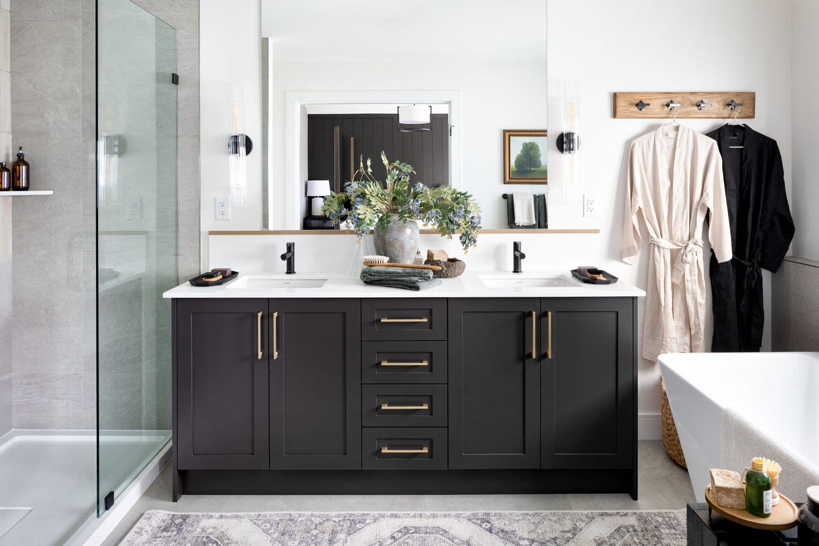 Mirada Estates – Primary bathroom with a dark charcoal double vanity, brass hardware, a walk-in glass shower, freestanding tub, and robes hanging on a wall hook.