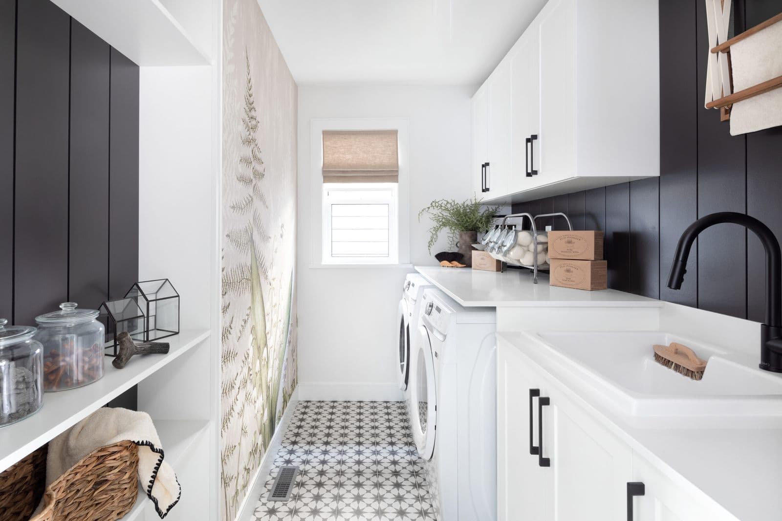 Mirada Estates – Narrow laundry room with white upper cabinets, black shiplap backsplash, a front-load washer and dryer, and patterned tile flooring.